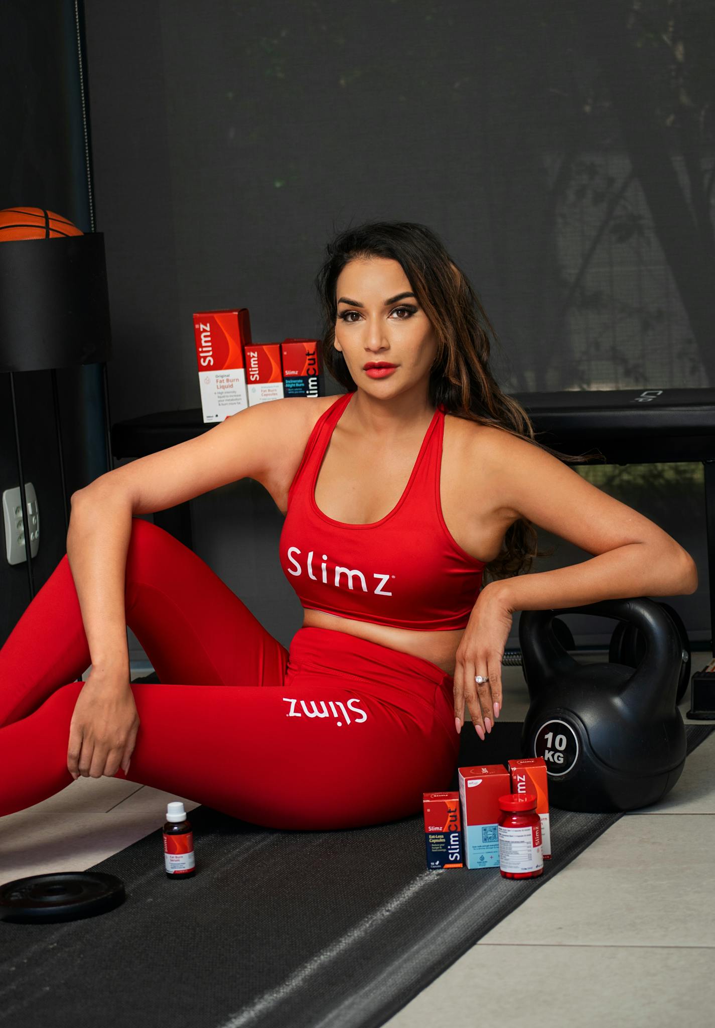 A woman in a red fitness outfit sits with nutrition supplements and a kettlebell indoors.
