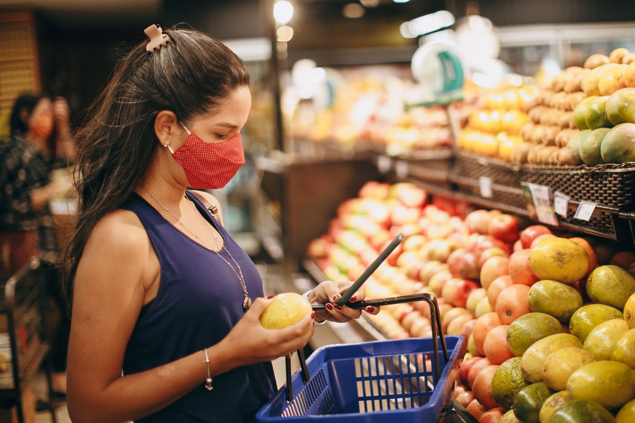 about-us-01 A woman wearing a mask selects fresh fruits in a supermarket produce aisle.
